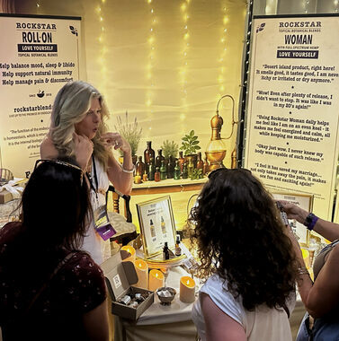 Heather Reilly Hiemstra gestures while laughing with two attendees at the Rockstar Blends booth, candles and botanical oils on the table, at Cannadelic Night Market.