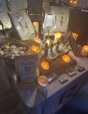A candlelit Rockstar Blends apothecary display—botanical oil bottles on a gold tray, framed product cards, a basket of botanical melts, and an email sign-up card—on the booth table at the Cannadelic Night Market.