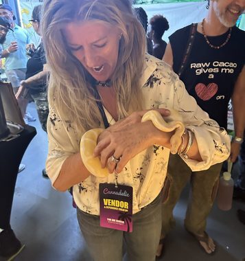 Heather Reilly Hiemstra smiles with delight while holding a ball python at the Cannadelic Night Market, wearing her Cannadelic vendor badge amid the festival crowd.