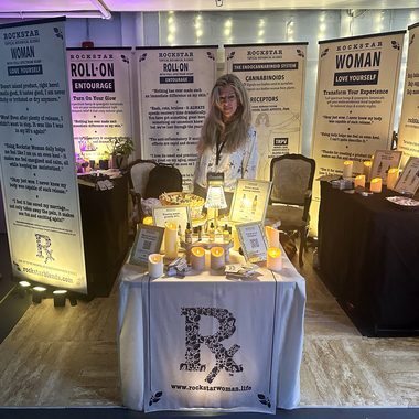 Heather Reilly Hiemstra, founder of Rockstar Blends, stands behind her candlelit botanical apothecary table—product trays and an "R" logo cloth reading rockstarwoman.life—framed by banners explaining the endocannabinoid system and the Rockstar Woman and Roll-On topicals at the Cannadelic Night Marke
