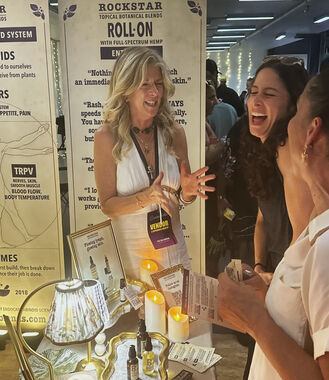 Heather Reilly Hiemstra, founder of Rockstar Blends, smiles with her arm around a guest in front of booth banners explaining the endocannabinoid system and the Rockstar Roll-On Entourage at Cannadelic Night Market.