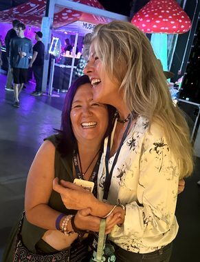 Heather Reilly Hiemstra laughs joyfully while embracing a longtime friend at the Cannadelic Night Market, a glowing red-and-white mushroom installation and string lights behind them.