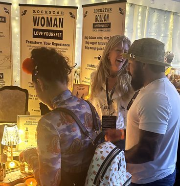 Heather Reilly Hiemstra laughs mid-conversation with two visitors at the Rockstar Blends booth, the candlelit table and Rockstar Woman and Roll-On banners behind her at the Cannadelic Night Market.