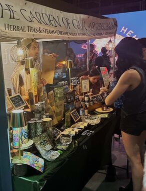 Heather Reilly Hiemstra laughs while holding a Rockstar Blends botanical oil at the booth, in front of the Rockstar Woman testimonial banner and the endocannabinoid-system diagram at Cannadelic Night Market.