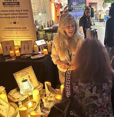 Heather Reilly Hiemstra greets a visitor with an animated smile at the Rockstar Blends booth, a neighboring apothecary's blue-lotus banner visible in the background at the Cannadelic Night Market.