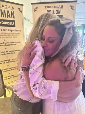 Heather Reilly Hiemstra shares a warm, eyes-closed embrace with a returning friend in front of the Rockstar Woman and Roll-On banners at the Rockstar Blends booth, Cannadelic Night Market.