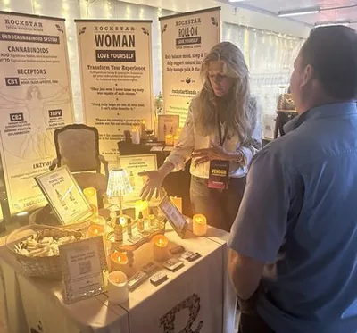 Heather Reilly Hiemstra gestures toward her product display while talking with a visitor at the Rockstar Blends booth, banners explaining the endocannabinoid system and an email sign-up card on the candlelit table, Cannadelic Night Market.