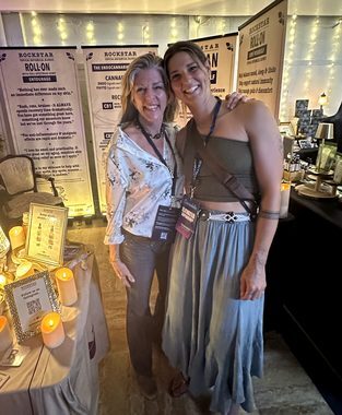 Heather Reilly Hiemstra poses with her arm around a guest at the Rockstar Blends booth, the candlelit apothecary table and endocannabinoid-system and product banners behind them at the Cannadelic Night Market.