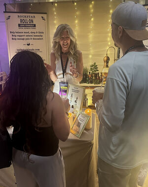 Heather Reilly Hiemstra speaks animatedly with two visitors holding drinks at the Rockstar Blends booth, the Roll-On "Love Yourself" banner, apothecary still, and an Instagram QR card behind her at Cannadelic Night Market.