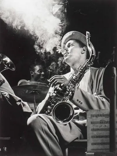 A saxophonist in a soft hat playing alto sax in a smoky club, a drummer behind