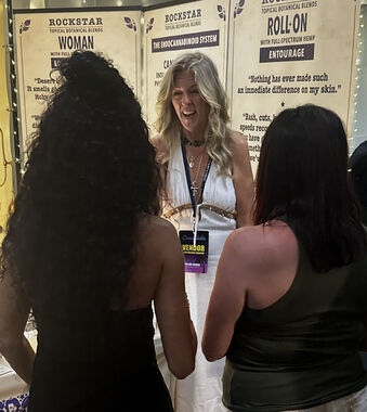 Heather Reilly Hiemstra speaks with two visitors at the Rockstar Blends booth in front of the Rockstar Woman, endocannabinoid-system, and Roll-On Entourage banners at Cannadelic Night Market.